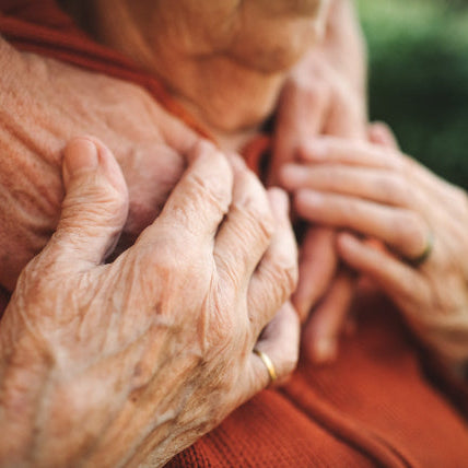 Close up shot of an elderly couple, holding hands on his chest as she stands behind him. He's wearing a red sweater, but you can not see either of their faces.
