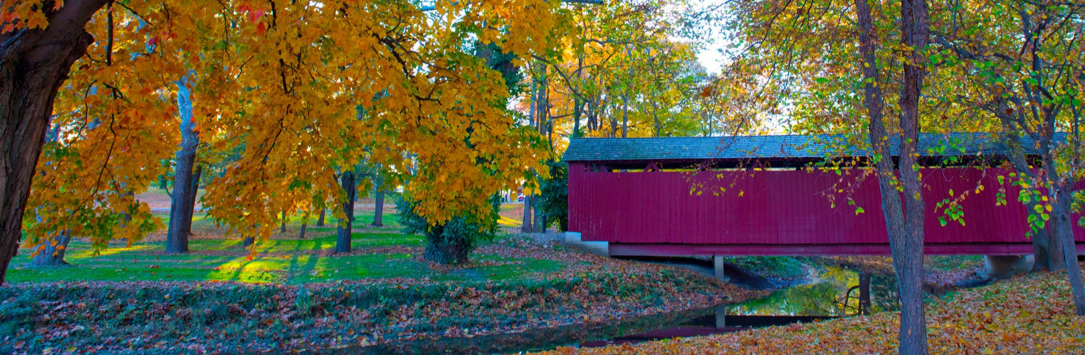 A red covered bridge spans a small creek in a wooded park during autumn in Indiana. The trees are full of vibrant fall foliage in shades of orange, yellow, and green. Fallen leaves scatter the ground, and the scene is calm and colorful with soft sunlight filtering through the trees.