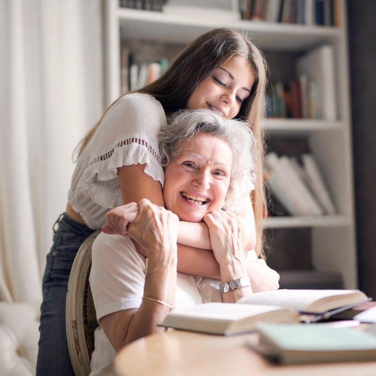 An elderly woman with gray hair sits at a table smiling brightly while being hugged from behind by a young woman with long brown hair. The young woman is also smiling and resting her head affectionately on the older woman's shoulder. There are open books on the table in front of them, and a bookshelf in the background.