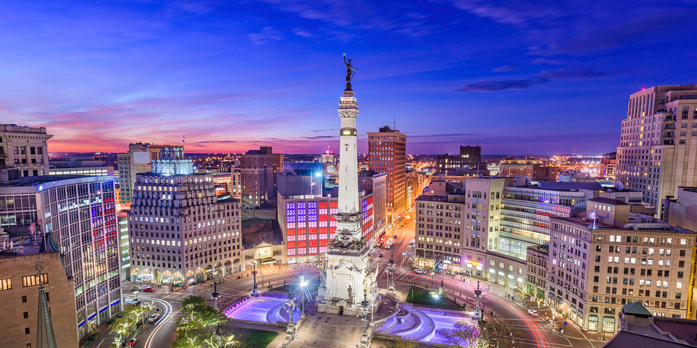 A vibrant evening view of downtown Indianapolis, Indiana, featuring the Soldiers and Sailors Monument at the center of Monument Circle. The monument is illuminated with white and purple lights, surrounded by historic and modern buildings. The sky is a gradient of blue, pink, and purple as the sun sets, and city lights begin to glow across the skyline.