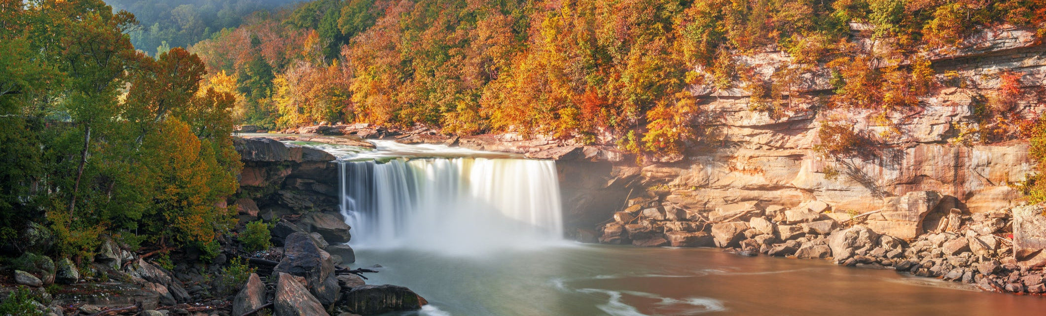 A wide waterfall cascades over a rocky ledge surrounded by vibrant autumn foliage in a forested area of Kentucky. The water flows into a calm river below, with large boulders scattered along the shoreline. The scene is bathed in warm sunlight, enhancing the rich colors of orange, red, and gold in the trees.