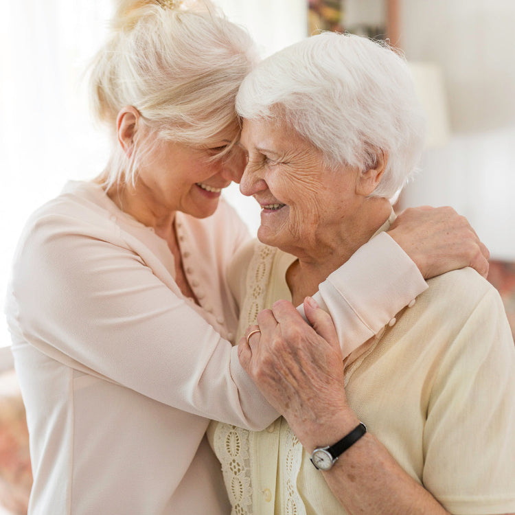 Two women share a warm embrace while smiling with their foreheads touching. One is an older woman with short white hair wearing a cream-colored blouse, and the other is a younger woman with blonde hair tied back, dressed in a light pink blouse. They appear joyful and close, standing in a softly lit indoor setting.