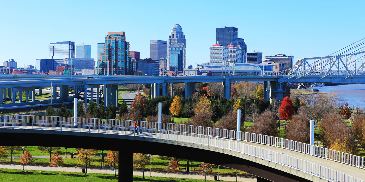 A view of downtown Louisville, Kentucky, featuring a mix of modern and historic skyscrapers under a clear blue sky. In the foreground, a curved pedestrian bridge winds through a park with autumn-colored trees. Behind the park, multiple layers of elevated highways lead toward the city's skyline. To the right, part of a bridge crosses the Ohio River.