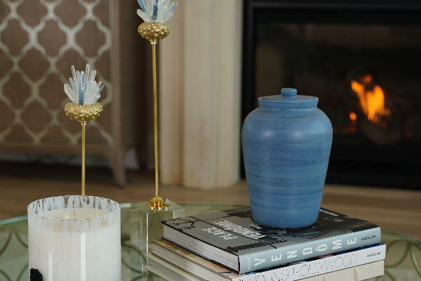 The Modern Classic Sky Memorial Urn by Stoneware & Co. is displayed in a warm, elegant home setting, resting atop a stack of designer books near a softly glowing fireplace. This handcrafted ceramic urn in serene sky blue features subtle variations in glaze, creating a peaceful and timeless tribute. Available through Magnolia Cremations, this urn blends contemporary design with meaningful remembrance.
