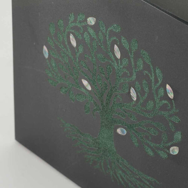 Close-up of a black marble cremation urn showing a detailed green inlay of a tree with swirling branches and roots, decorated with iridescent, leaf-shaped accents.
