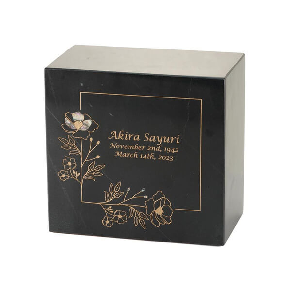 Black marble cremation urn with gold floral line art and a decorative inlay of one iridescent flower; optional engraving is shown with name and dates.