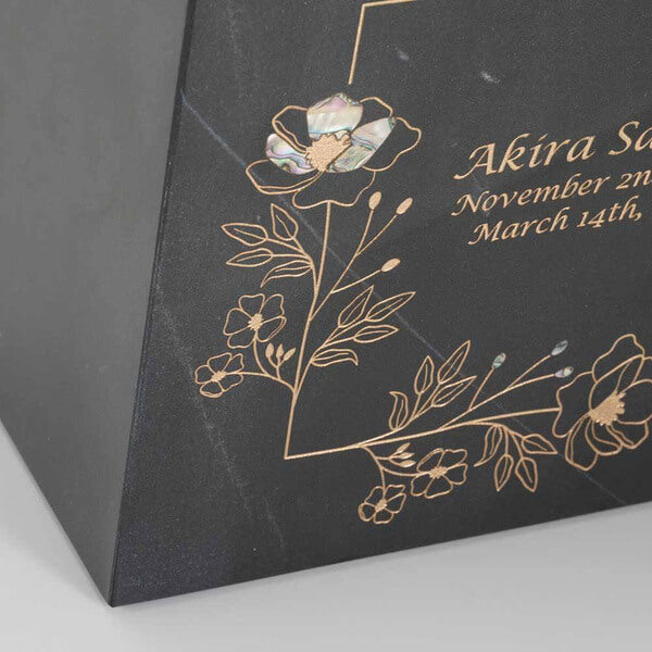 Close-up of a black marble cremation urn featuring gold floral line art with one flower inlaid with iridescent material; partial view of engraved name and dates is also visible.
