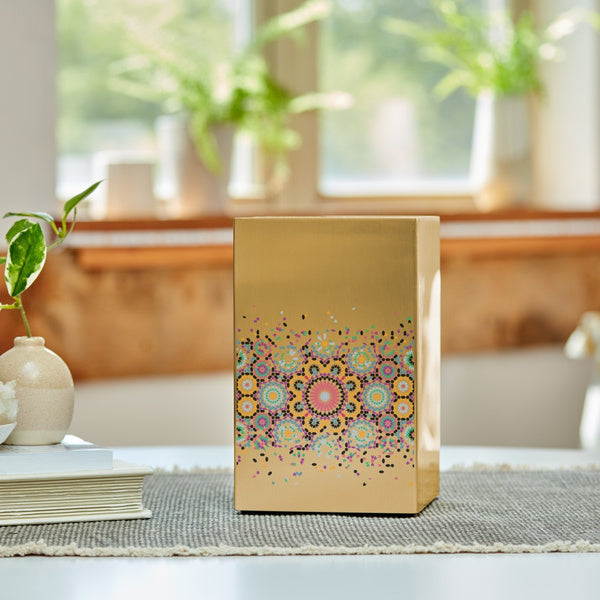 The Mosaic in Bronze Memorial Urn from Magnolia Cremations sits gracefully on a textured placemat, illuminated by natural light. This contemporary cremation urn, crafted from brushed stainless steel with a bronze-tone finish, features a vibrant mosaic-inspired design with intricate circular patterns and scattered confetti-like accents. A meaningful and artistic tribute, blending elegance with a heartfelt celebration of life.