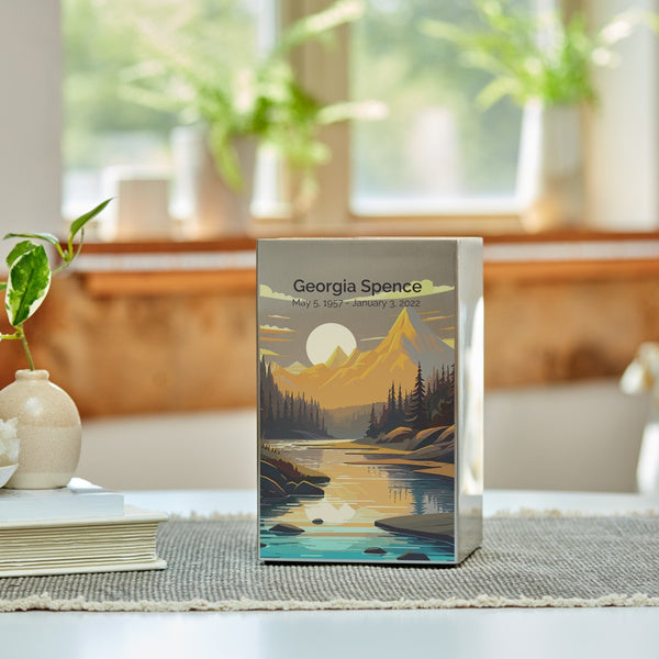 The Mountain Landscape in Pewter Memorial Urn by Magnolia Cremations is showcased on a softly lit table, with natural light filtering through a nearby window. The urn's brushed pewter finish features a serene mountain scene with a glowing sun setting behind rugged peaks, reflecting on a tranquil river surrounded by evergreen trees. A potted plant and neutral-toned décor enhance the peaceful ambiance. Engraving is shown as an available option, allowing for a personalized tribute.