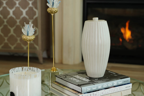 The Nouveau Groove Beach Memorial Urn by Stoneware & Co. is displayed on a glass coffee table, resting atop a stack of hardcover books. The urn’s elegant, fluted design in soft beach beige complements the warm, modern setting, which includes a flickering fireplace, a large candle, and decorative gold and glass ornaments. The scene evokes a sense of tranquility, offering a graceful tribute to a loved one.
