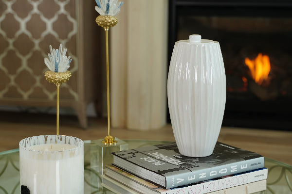 A full-size ceramic urn in cloud white from the exclusive Nouveau Groove collection for Magnolia Cremations by Stoneware & Co. The urn has an elegant Art Nouveau-inspired design, featuring vertical ridges and a smooth, rounded lid. It is displayed in a home setting on a glass coffee table, resting atop a stack of designer books. A softly glowing fireplace and decorative elements, including a white candle and gold-accented sculptures, create a warm and peaceful ambiance in the background.