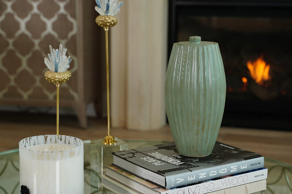 A full-size ceramic urn in forest green from the exclusive Nouveau Groove collection for Magnolia Cremations by Stoneware & Co. The urn has a gently tapered shape with vertical ridges, giving it an elegant Art Nouveau-inspired design. It sits atop a stack of books on a glass coffee table in a cozy living room setting, with a fireplace glowing in the background. Nearby, decorative accents, including a white candle and gold-and-glass ornaments, enhance the serene ambiance.