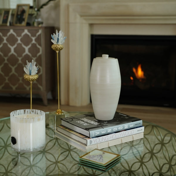 A full-size ceramic urn in beach beige from the exclusive Nouveau Pure collection for Magnolia Cremations by Stoneware & Co., displayed on a glass coffee table. The urn has a smooth, tapered shape with a minimalist design, complementing the modern decor of the living space. It is positioned on top of stacked hardcover books, with a candle, decorative gold and blue floral sculptures, and a fireplace in the background, creating a serene and elegant setting.