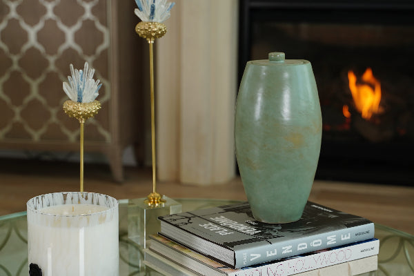 The Nouveau Pure Forest Memorial Urn, an exclusive design for Magnolia Cremations by Stoneware & Co., is displayed on a glass coffee table, resting atop a stack of designer books. This full-size ceramic urn has a smooth, tapered silhouette with a tranquil forest green glaze. The setting features a softly lit living space with elegant decor, including a burning fireplace in the background, decorative gold and glass floral accents, and a white marbled candle. The urn’s design inspired by Art Nouveau.