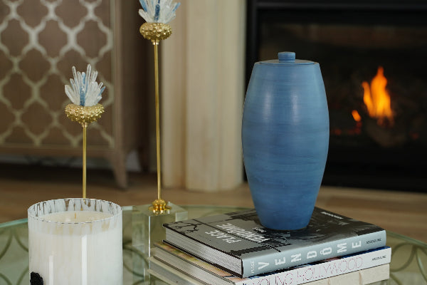 The Nouveau Pure Sky Memorial Urn, an exclusive design for Magnolia Cremations by Stoneware & Co., is elegantly displayed atop a stack of books on a glass coffee table. The urn is a full-size ceramic piece with a smooth, tapered silhouette and a serene sky blue glaze, featuring subtle tonal variations. In the softly lit background, a fireplace casts a warm glow, complementing the urn’s calming presence.