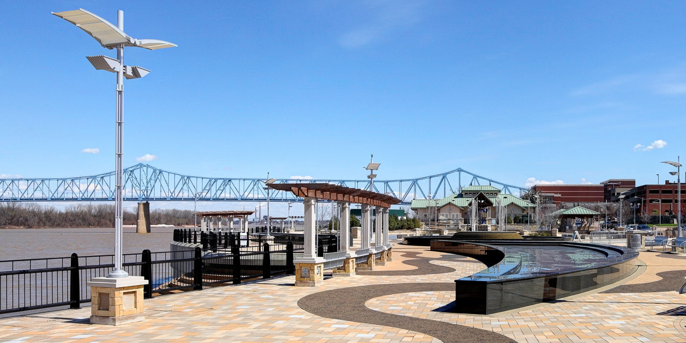 A modern riverfront park in Owensboro, Kentucky, featuring a wide walkway with decorative stone paving, curved black railing, and a large water feature. A pergola-covered seating area runs alongside the Ohio River. In the background, a blue truss bridge spans the river, and buildings with green roofs are visible under a clear blue sky.