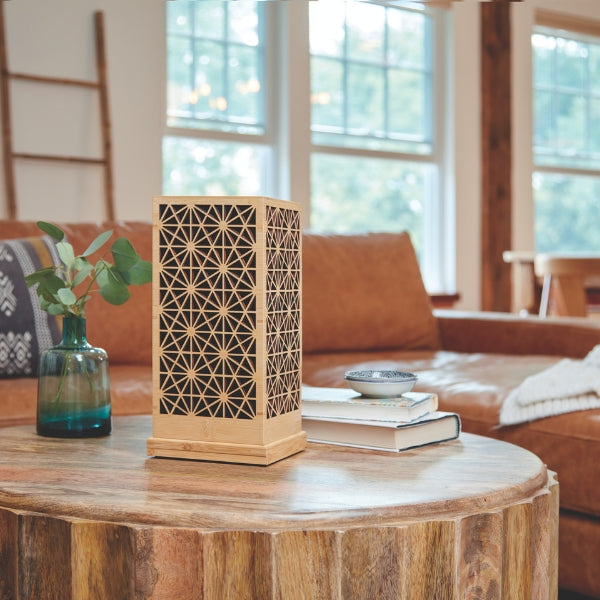 The Palazzo memorial urn, available at Magnolia Cremations, sits elegantly on a rustic wooden coffee table in a cozy living space. The urn is crafted from bamboo and black MDF, featuring an intricate geometric lattice pattern on all visible sides. The setting includes a brown leather couch, a stack of books, a small decorative bowl, and a glass vase with greenery, creating a warm and inviting atmosphere.
