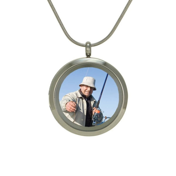 The Pewter Round Photo Glass Pendant Necklace, available at Magnolia Cremations, is shown with a sample photo inside the hinged glass front. This stainless steel cremation pendant allows for easy photo insertion, combining a cherished image with a small portion of ashes for a deeply personal keepsake. The design includes engraving options for customization and comes with a matching 19-inch chain and a lobster claw clasp.