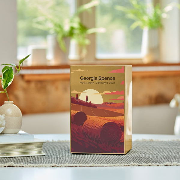 A Prairie Landscape in Bronze Memorial Urn sits on a neutral woven mat in a sunlit room, available at Magnolia Cremations. The urn features a warm-toned illustration of a prairie at sunset, with rolling hills, hay bales, and a golden sun. A personalized engraving is displayed on the brushed bronze surface, highlighting the option for customization. The urn is placed in a peaceful setting with soft natural light, green plants, and a ceramic vase, creating a comforting tribute.