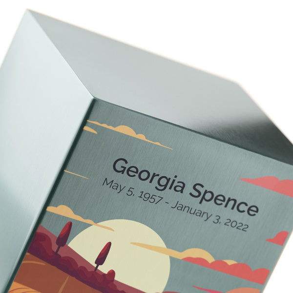 A close-up of the Prairie Landscape in Pewter Memorial Urn, available at Magnolia Cremations, highlighting its brushed stainless steel surface and personalized engraving. The urn features a serene prairie landscape design with a warm sunset, rolling hills, and silhouetted trees. The smooth metallic finish reflects soft light, emphasizing the craftsmanship and engraving options available for customization.