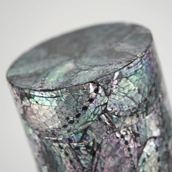 Close-up view of the top portion of a cylindrical scattering urn, featuring a glossy, iridescent surface with abalone shell pieces arranged in a mosaic floral pattern. The surface shows multicolored hues of green, purple, and silver with visible texture and cracks.