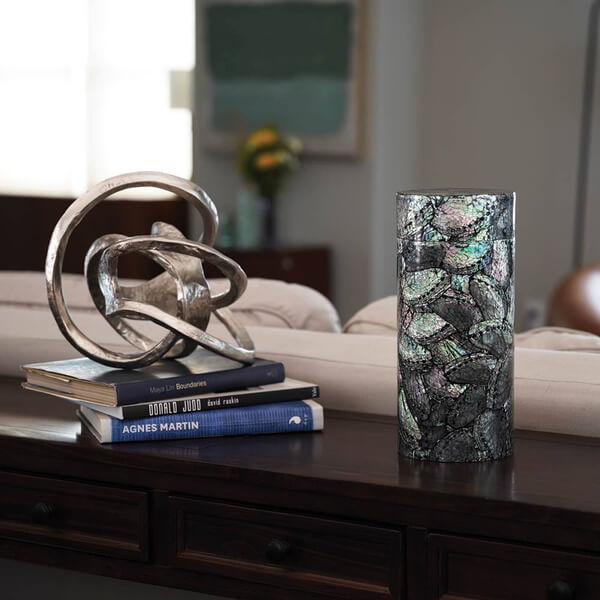 Cylindrical scattering urn with an iridescent abalone shell floral mosaic design displayed on a dark wooden table in a living room. The urn is placed beside a decorative metallic sculpture resting on stacked art books, with a neutral-colored couch and abstract wall art visible in the background.