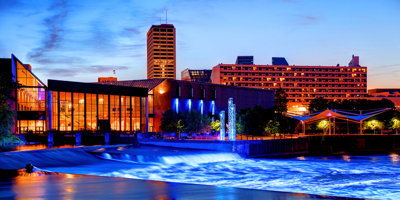 Evening view of downtown South Bend, Indiana, featuring the Century Center convention complex with its large glass windows illuminated in warm orange light. In the foreground, the St. Joseph River flows over a low dam, lit with vibrant blue lights. Modern sculptures and a white canopy structure are visible along the riverside park, with tall buildings and a deep blue sky in the background.