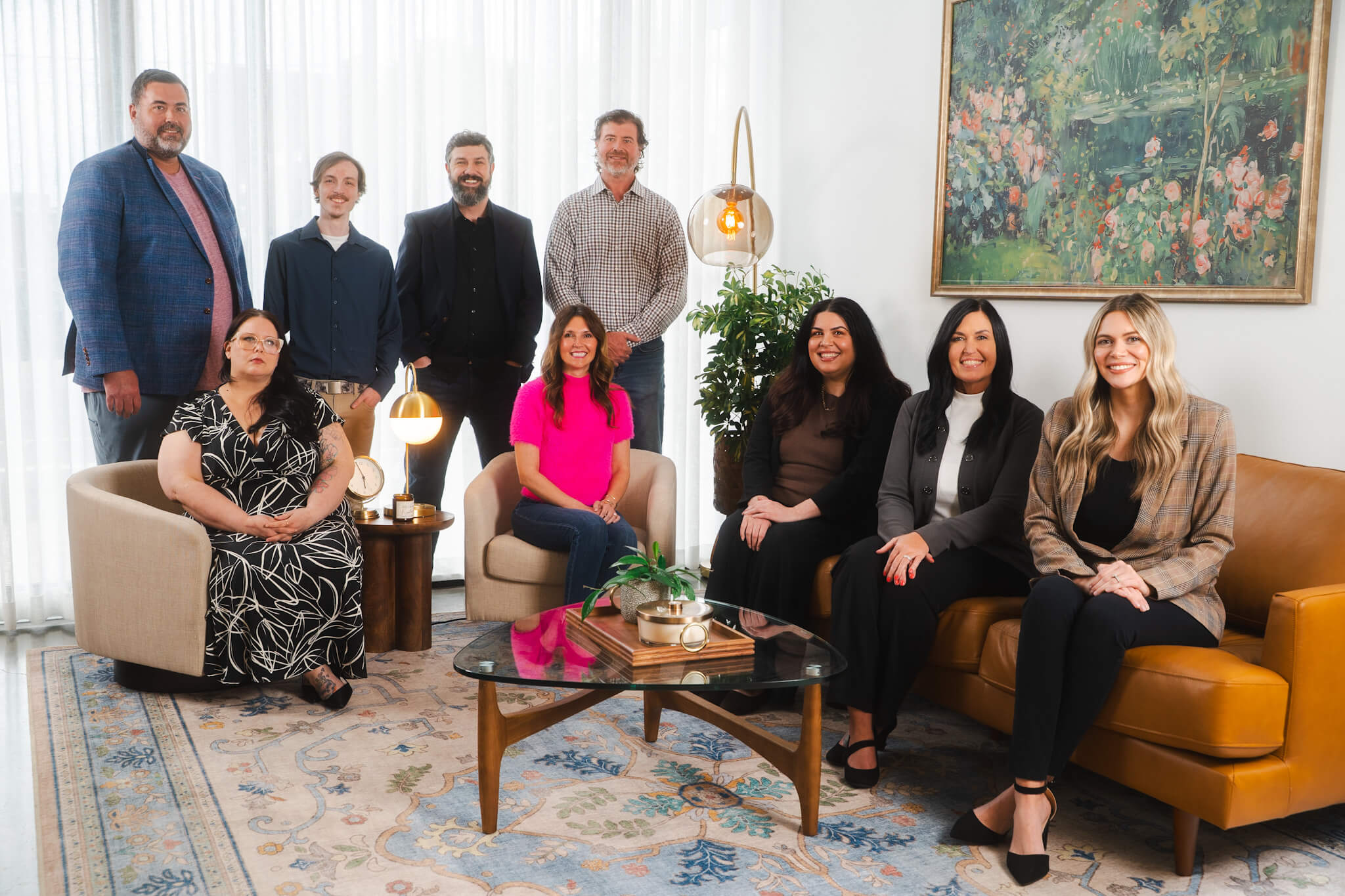Group portrait of the Magnolia Cremations team. Four men stand in the back row from left to right: Aaron, Brandon, Jason, and Billy. Five women sit in the front row from left to right: Princessa in a black and white dress, Mandi in a bright pink top, Carolina in a brown top, Shanna in a gray jacket, and Abby in a plaid blazer. The team is posed in a warmly lit, modern office space with mid-century-style furniture, a large floral painting on the wall, and a patterned rug beneath them.