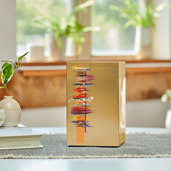 The Artist in Bronze Memorial Urn, available at Magnolia Cremations, is displayed on a textured gray table runner in a softly lit room. This contemporary stainless steel urn features a striking abstract design with vibrant colors and expressive brushstrokes, creating a modern and artistic tribute. Natural light from the window enhances the urn’s brushed bronze finish, complementing the serene and elegant setting.