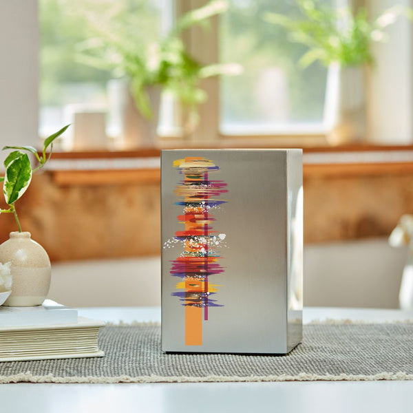 The Artist in Pewter Memorial Urn, available at Magnolia Cremations, sits on a neutral-toned table runner in a bright, sunlit home. The brushed stainless steel urn features a striking, colorful abstract design, adding a modern and artistic touch. Green plants and soft natural light in the background create a peaceful and reflective atmosphere.