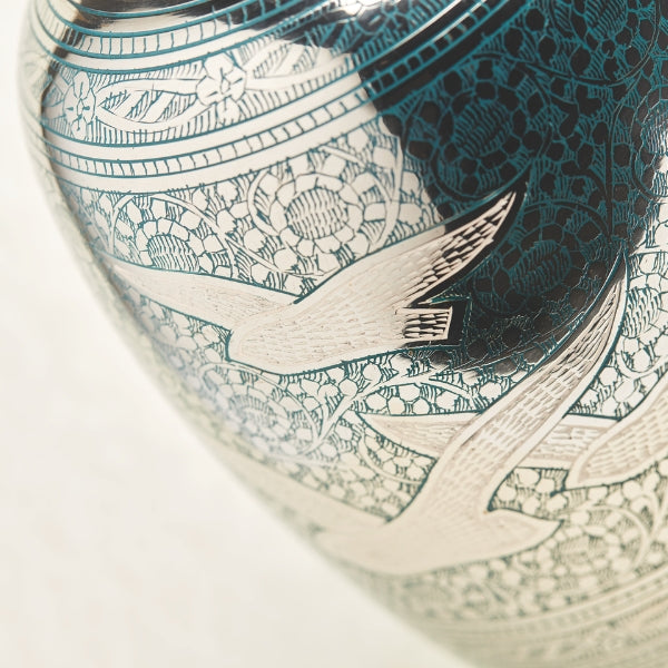 A close-up view of the Traditional Going Home Memorial Urn, available at Magnolia Cremations, showcasing its intricate hand-engraved design. The reflective silver and green nickel-plated brass surface features a delicate floral pattern with three gracefully soaring birds. The fine detailing highlights the craftsmanship and elegance of this meaningful tribute.