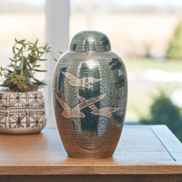 The Traditional Going Home Memorial Urn, available at Magnolia Cremations, sits on a wooden surface near a window, with natural light reflecting off its polished nickel-plated brass finish. The urn features an intricate silver and green floral engraving with three gracefully soaring birds. A matching engraved lid completes the elegant design, creating a timeless and meaningful tribute. A potted plant and a blurred outdoor landscape are visible in the background.