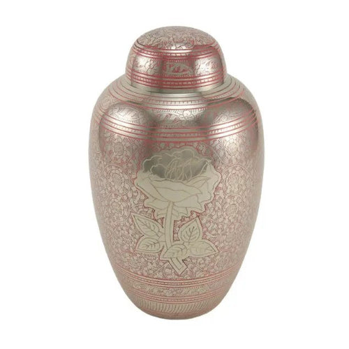 The Traditional Rose Memorial Urn, available at Magnolia Cremations, is a full-size brass urn with a reflective pink and silver finish. Its surface features an intricate floral engraving, with a beautifully detailed silver rose at the center. The urn's delicate hand-engraved patterns and timeless design offer a meaningful tribute to a loved one.