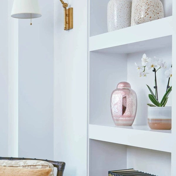 The Traditional Rose Memorial Urn, available at Magnolia Cremations, is displayed on a white built-in shelf alongside a potted orchid and ceramic vases. The urn features a reflective pink and silver finish with intricate floral engravings and a central pewter rose design, offering a timeless and elegant tribute.
