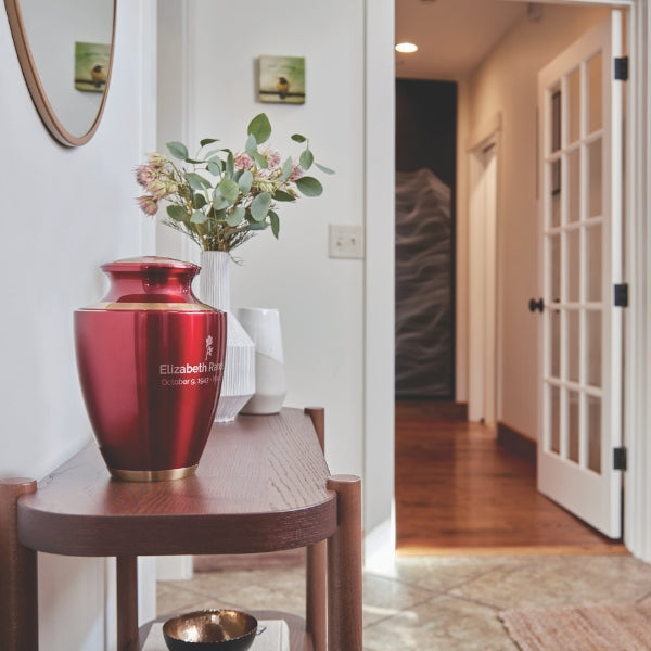 The Trinity Crimson Memorial Urn, available at Magnolia Cremations, displayed on a wooden console table in a well-lit home entryway. The urn features a deep red high-gloss finish with gold accents and an engraved personalization on the front. A vase with fresh flowers and decorative ceramics sit nearby, creating a peaceful and elegant memorial setting.