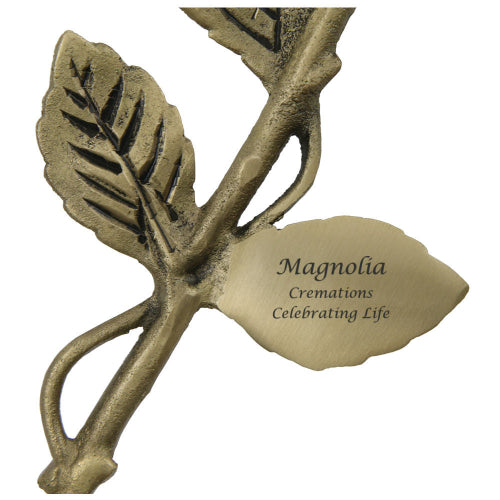This image shows a close-up of the White Rose Keepsake from Magnolia Cremations, a brass memorial piece shaped like a delicate rose. The golden-toned stem features textured leaves and an engraved plaque with a smooth, polished surface. Designed to hold a small portion of ashes, this elegant keepsake serves as a lasting tribute to a loved one, blending artistry with remembrance.