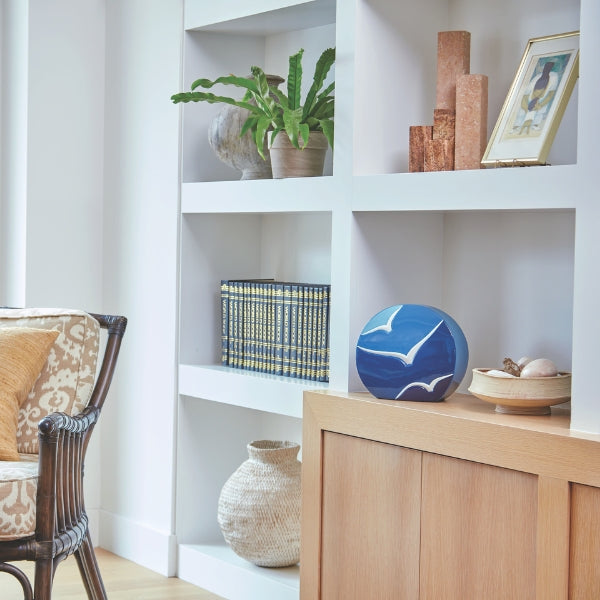 The Windham Going Home Glossy Blue Memorial Urn, available at Magnolia Cremations, is displayed on a light wood cabinet in a modern living space. The urn's curved, sculptural form features a glossy blue gradient, with three white birds in relief soaring across its surface. Its contemporary design allows it to blend seamlessly with home décor, surrounded by neutral tones, woven baskets, lush greenery, and carefully curated decorative pieces. Positioned on an open shelf, the urn serves as a meaningful tribute