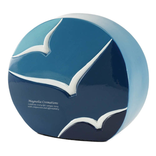 The Windham Going Home Glossy Blue Memorial Urn, available at Magnolia Cremations, is a contemporary ceramic urn with a unique rounded shape. The urn features a smooth, glossy gradient transitioning from deep navy at the base to a lighter sky blue at the top. Three white bird silhouettes, sculpted in relief, soar across the surface, symbolizing peace, freedom, and the journey home. An engraving is displayed on the lower left side, showcasing available personalization options.