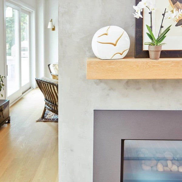 The Windham Going Home Glossy White Memorial Urn, available at Magnolia Cremations, is displayed on a wooden mantel above a modern fireplace. The urn has a unique curved shape with a glossy white surface and hand-painted gold birds in flight, symbolizing peace and serenity. The surrounding decor includes a potted white orchid, a framed artwork, and a minimalist interior with soft natural lighting.