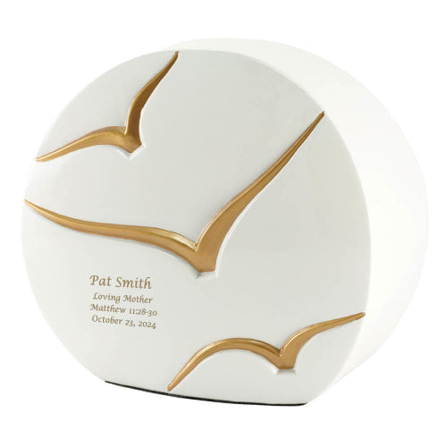 The Windham Going Home Glossy White Memorial Urn, available at Magnolia Cremations, features a contemporary curved design with a glossy white finish and three gold-painted birds in flight, symbolizing peace and freedom. The urn's smooth ceramic surface and elegant detailing make it a refined tribute for a loved one. Engraving is shown as an available option.