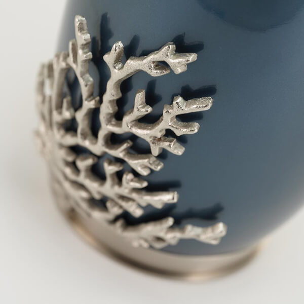 Close-up of the Windsong Coral Memorial Urn, available at Magnolia Cremations. The image highlights the detailed pewter coral accent, which extends from the base of the urn. The textured coral design contrasts beautifully with the smooth, deep blue high-gloss finish of the stainless steel body. A subtle brass accent at the base adds a refined touch.