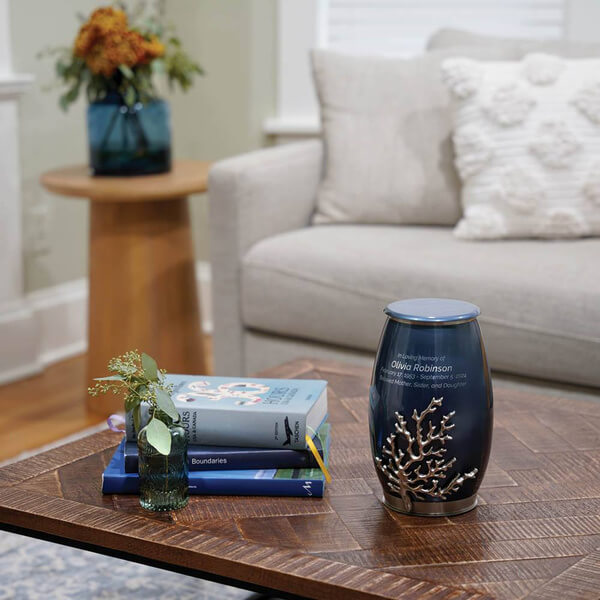 The Windsong Coral Memorial Urn, available at Magnolia Cremations, displayed on a wooden coffee table in a cozy living room. The urn features a deep blue high-gloss finish with a detailed silver coral accent extending from the base. A soft engraving is visible on its smooth surface. Beside the urn, a small green glass vase with sprigs of greenery and a neatly stacked selection of books add to the serene ambiance. A neutral-toned sofa with textured pillows and a side table with a floral arrangement complete