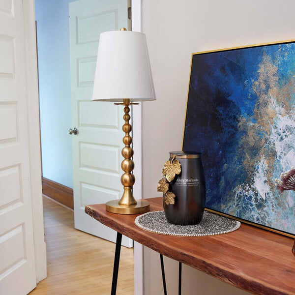 Slate gray cremation urn with gold leaf vine decoration displayed on a wooden table in a home interior. The urn sits on a decorative mat next to a gold and white table lamp and a blue and gold abstract painting. Optional engraving is shown on the urn.