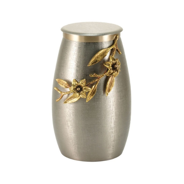 The Windsong Lily Pewter/Bronze Memorial Urn, available at Magnolia Cremations, is an artistic stainless steel urn with a gently tapered shape and a brushed pewter finish. A sculpted bronze-finished lily branch wraps around the upper portion, featuring delicate flowers and leaves in high relief. The flat, round lid is accented with a subtle gold trim, complementing the floral embellishment.