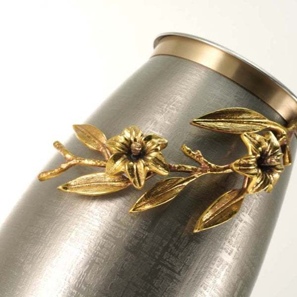 A close-up view of the Windsong Lily Pewter/Bronze Memorial Urn, available at Magnolia Cremations. The urn’s brushed pewter finish has a subtle texture, complementing the intricate gold-toned floral embellishment. The raised bronze-finished lily branch extends across the surface, showcasing delicate flowers, leaves, and buds. The flat lid features a coordinating gold accent, adding a refined touch to this artistic design.