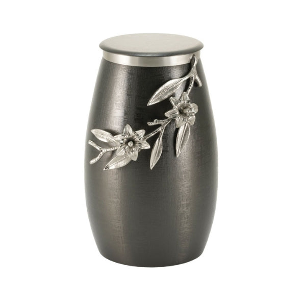 The Windsong Lily Slate/Pewter Memorial Urn, available at Magnolia Cremations. This urn features a sleek, tapered silhouette with a deep slate-black brushed finish, adding a contemporary elegance. A sculpted pewter lily branch with delicate flowers and leaves extends across the urn, creating a striking contrast against the dark background. The flat stainless steel lid is accented with a silver rim, complementing the detailed floral embellishment.
