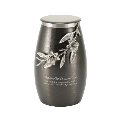 he Windsong Lily Slate/Pewter Memorial Urn, available at Magnolia Cremations. This urn features a brushed slate-black stainless steel finish with a sculpted pewter lily branch wrapping around the side, showcasing delicate flowers and leaves in high-relief detail. The urn has a contemporary, tapered shape, and the flat stainless steel lid complements the pewter floral embellishment. An engraving is displayed, indicating the option for personalization.