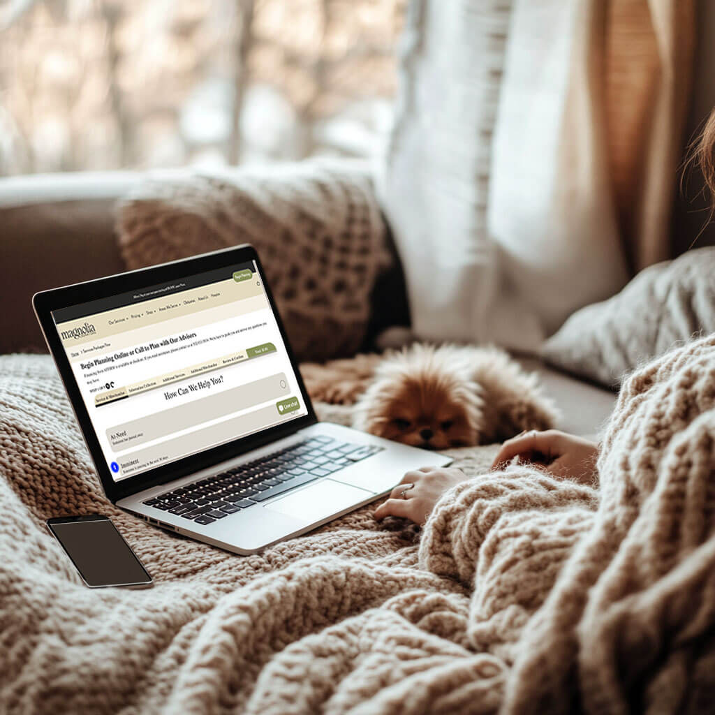 A person comfortably wrapped in a cozy knit blanket, using a laptop to plan a cremation online through Magnolia Cremations' easy-to-use website. A small dog rests nearby, adding to the warm and stress-free experience.