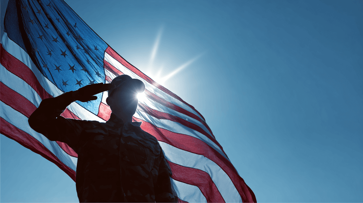 Silhouetted figure of a U.S. soldier in uniform saluting in front of a large American flag, with the sun shining behind their head against a clear blue sky.
