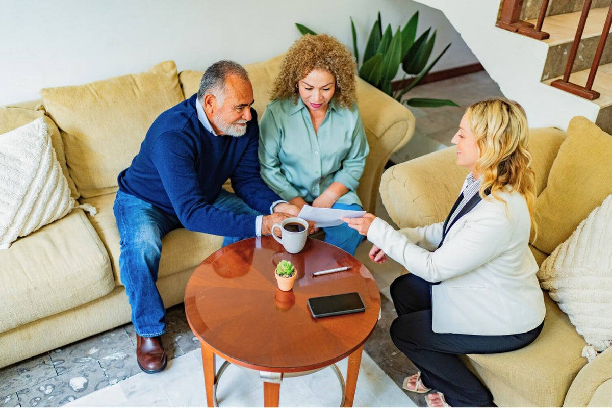 Three adults sitting in a living room having a discussion. An older couple sits on a beige couch reviewing a document handed to them by a woman in professional attire. A coffee table between them holds a coffee mug, smartphone, pen, and small plant.